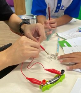 Teacher demonstrating electromagnetism with a battery circuit and paper clips.